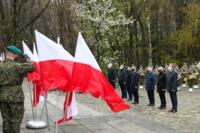 Na zdjęciu powiewają flagi polski, obok stoją żołnierze w mundurach a w tle widać stojących na baczność ludzi