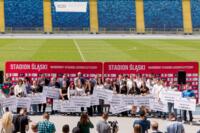 ludzie stojący w szeregu z czekami o dużych kwotach na stadionie w słoneczną pogodę