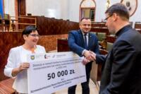 Po lewej szczęśliwa kobieta trzyma czek na 50 tysięcy złoty. Po jej prawej stronie stoi mężczyzna w garniturze, który dostaje gratulacje od innego mężczyzny w garniturze stojący przed nimi