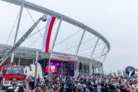 Po lewej jest stadion z wiszącą na nim flagą Polski. Pod nim bardzo duża ilość osób.