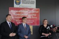 Z lewej strony stoi dwóch mężczyzn w garniturach i słuchają co mówi kobieta stojąca po prawej stronie. Kobieta elegancko ubrana odpowiada na pytania. Ma przed sobą masę mikrofonów